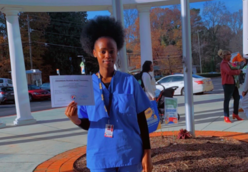Sandrine standing outside Georgia Piedmont Technical College wearing blue nursing scrubs holding her Nursing Assistant Certification.