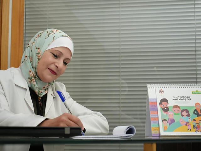 A woman wearing a head wrap and smiling sits at a table, writing with a pad and pen.