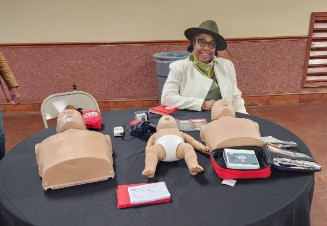 Stacy Robinson sitting at a table with her CPR training supplies, including two adult CPR dummies and one baby CPR dummy, laid out in front of her.