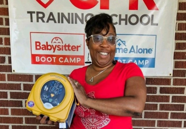 Stacy in front of a CPR Training school sign holding up a defibrillator.