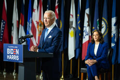 President Joe Biden stands at a podium giving a speech while Vice President Kamala Harris sits on stage. The new administration takes office after four years of Trump Administration policies that wreaked havoc on the lives of refugees and asylum seekers. 