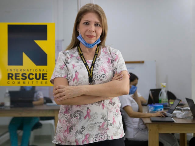 Dr. Edna Patricia Gomez stands in front of an IRC logo in a clinic wearing her medical scrubs and a mask around her chin. 