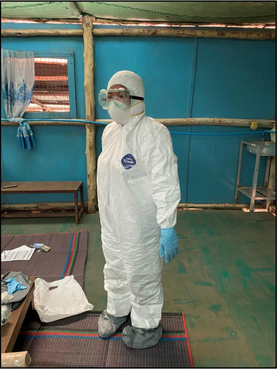 A refugee medic stands inside in a room. They are wearing full personal protective equipment, or PPE, including a white suit, blue gloves, and goggles.