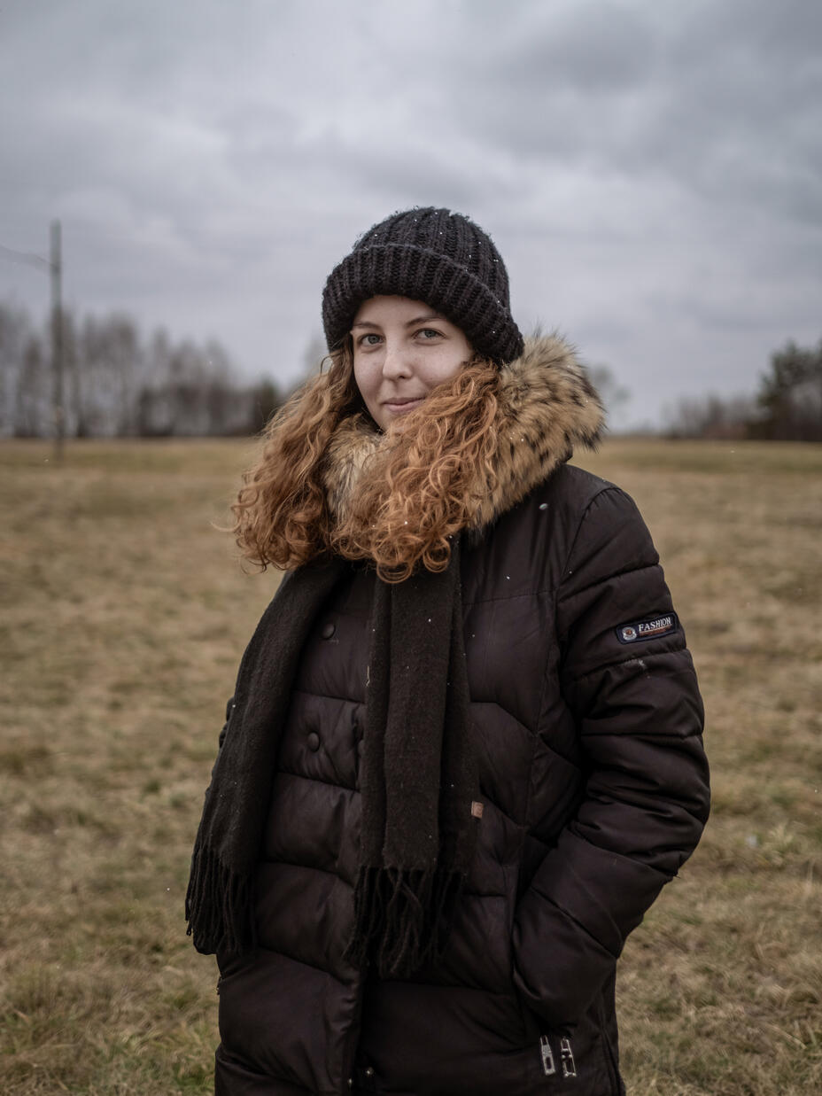 A young woman wearing a winter hat and jacket stands in a field and looks directly in the camera. 