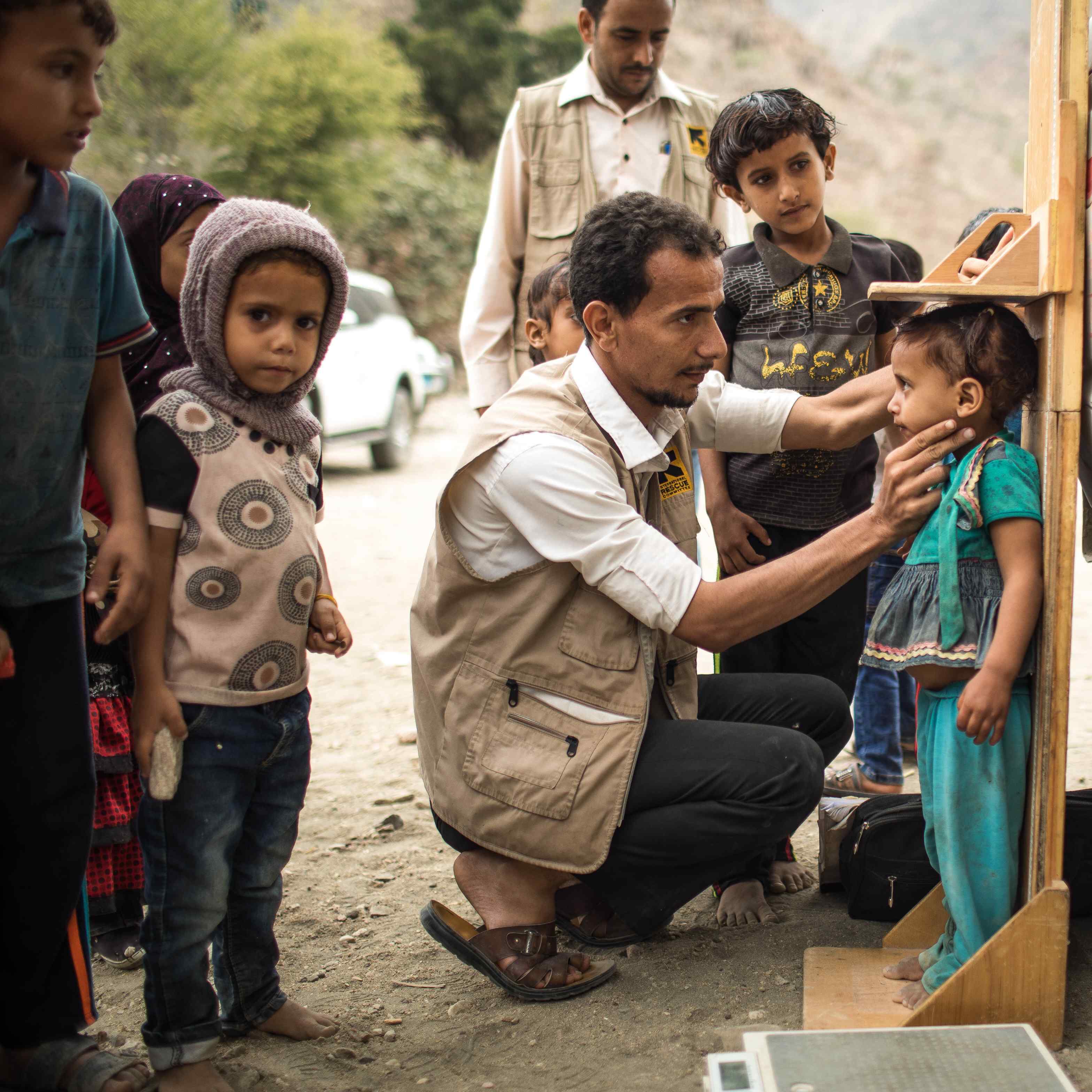 IRC 이동진료소의한남성의료진이예멘의외딴산지마을에서영양실조 징후를보이는아동을진찰하고, 다른아동들이그장면을지켜보고있습니다.