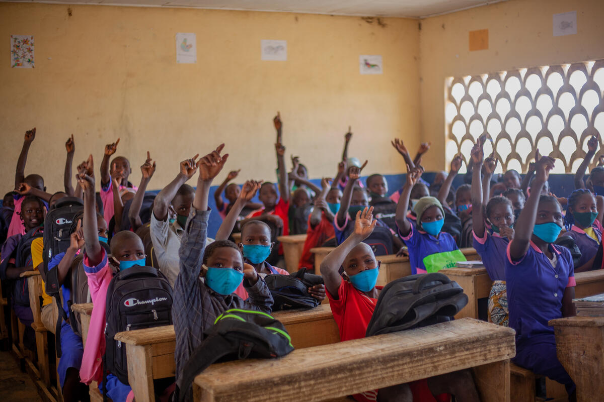 Children seated at wooden desks raise their hands in a classroom with breeze block windows in northern Cameroon. 