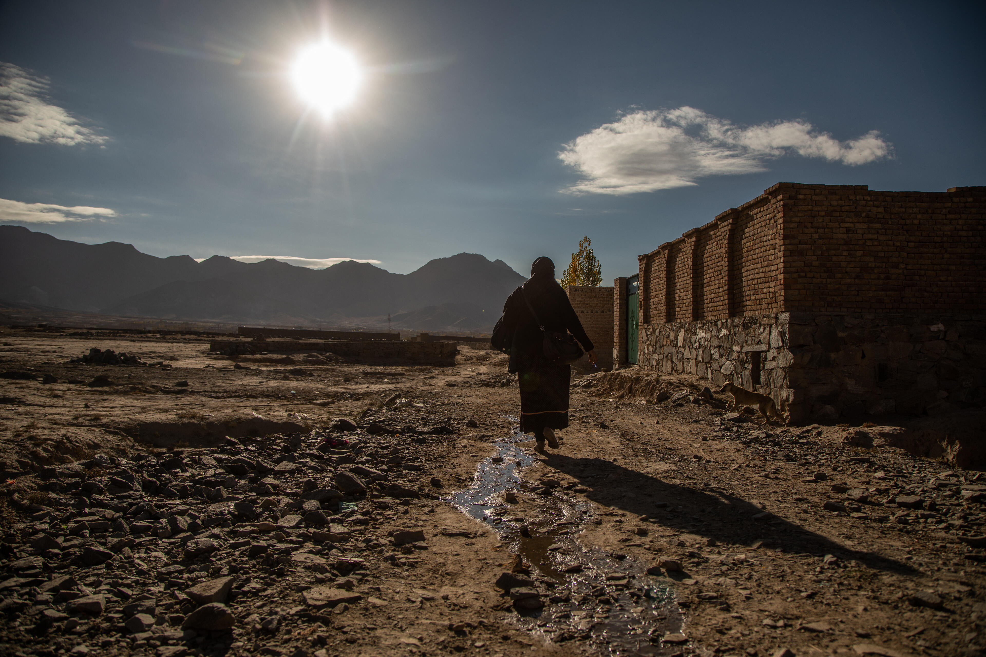 An Afghan aid worker is silhouetted against bright sunlight as she walks to a home in a village in a rocky landscape.