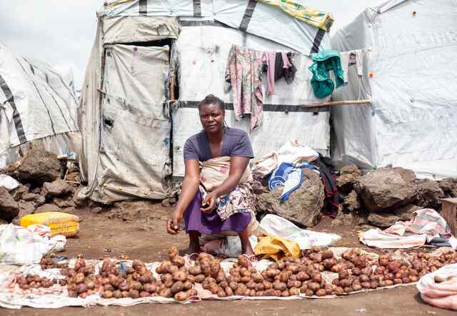 En kvinna sitter ner på marken framför ett vitt tält, hon säljer potatis. 