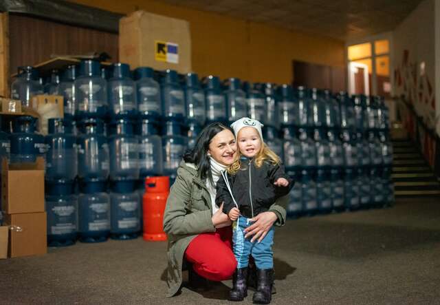 A mother and a daughter smile while posing for a photo in front of a room full of empty gas canisters that are to be filled and distributed to Ukrainians in need.