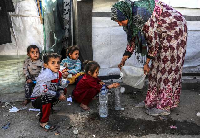 A woman pours water into a young girl's water bottle. Three other children sit nearby, watching on.