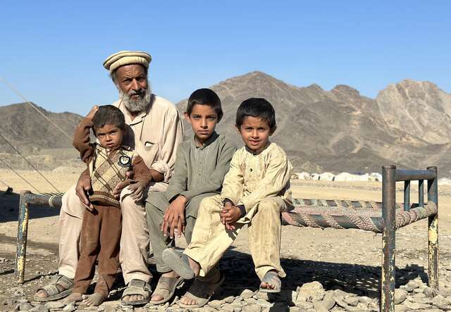 A man and his three sons pose for a photo, while sitting on a cot in the desert.