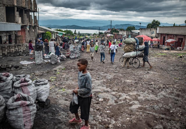A young boy and other people on the streets of Goma under a cloudy sky with mountains and a lake in the background.