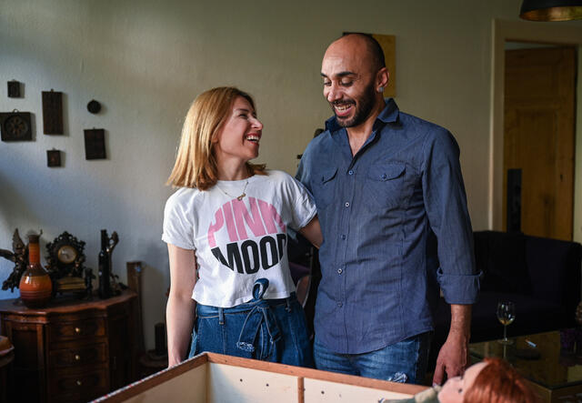 Bashar, wearing a blue button-down shirt, and his wife Lama, wearing a white t-shirt, stand with their arms around one another. They both have big smiles on their faces and are looking at one another. 