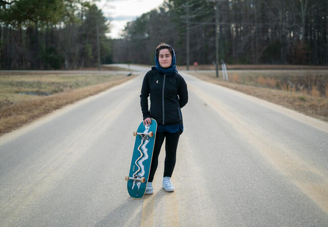 Belqisa stands on a street with a forest in the background. She is holding up her skateboard and looking at the camera 