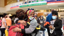 A refugee family is reunited at an American airport. The family hug each other in a warm embrace and smile.