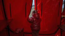 A young girl wearing a red winter coat stands in a red tent, peering out.