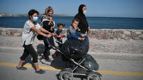 Asylum seeker mothers and their young children walk down the road in Lesvos with the sea in the background. 