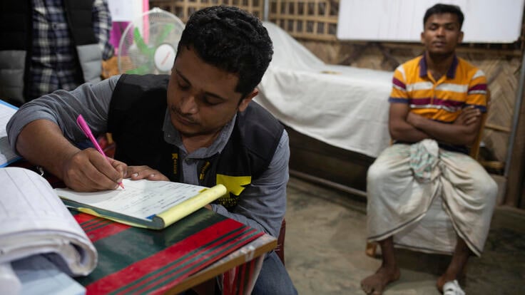Dr. Mahmudul Hossain, wearing an IRC vest, sits at a desk writing notes. A patient sits behind him. 