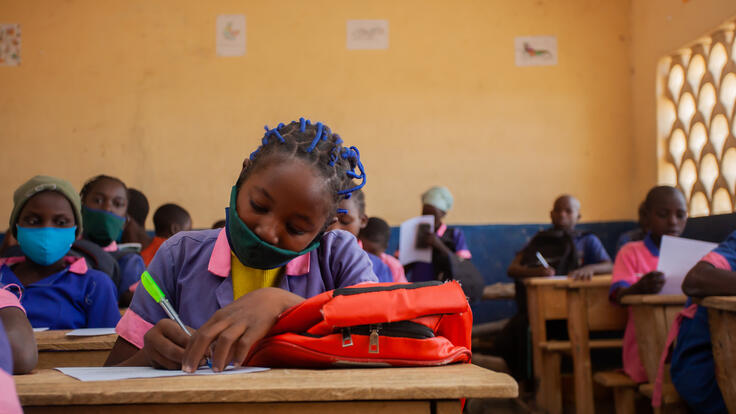 Eleven-year-old Kauvaumah, wearing her school uniform, writes at a wooden desk in her classroom in Cameroon. 