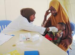 소말리아의 국제구조위원회 의료 종사자가 테이블에 기대어 엄마 무릎에 있는 아기를 검사하고 있습니다.