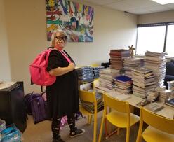 An IRC staff wearing a pink backpack and standing in front of a large pile of donated notebooks and other school supplies.