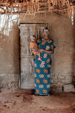 A mother poses for a portrait while holding a young child in her arms. Both look at the camera while standing on a dirt floor in front of their home in Mali.