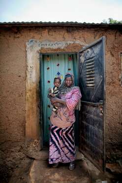A mother, wearing  a pink outfit, holds her daughter on her hip outside of their home.