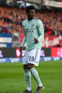 Alphonso Davies im grünem Trikot des FC Bayern München steht auf dem Spielfeld und zeigt mit dem Daumen nach oben.