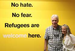Jeff and Mary stand side by side to the right of the photo, arms wrapped behind each other, in front of an IRC yellow wall and a wall that reads: "No hate. No fear. Refugees are welcome here."