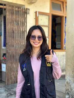 A young woman with long brown hair and sunglasses is looking at the camera and doing a thumbs up with her left hand