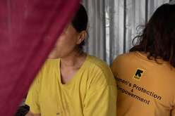 Nwe and another woman are back to back, they both are wearing yellow t-shirts. We see the logo of the IRC with "Women's protection and empowerment".