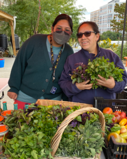 A woman in glasses and a mask stands close to her mother, a smiling woman with sunglasses on, in front of fresh vegetables at a farm stand. 