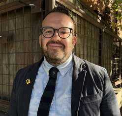 Headshot of Danny Beus, executive director at the International Rescue Committee in Utah, dressed in a grey suit & tie, light blue button up, IRC pin, and glasses. 