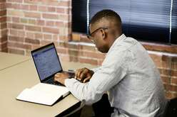 Man with glasses sits facing a laptop at a desk with an open notebook to the side. He is dressed in a long-sleeved button up with glasses. Red brick wall and window with the blinds closed & down in the background. 