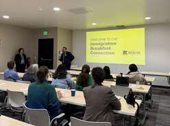 Classroom photo taken behind the students with presenters facing the camera; multiple students seated at tables looking toward two presenters standing with a large IRC graphic behind welcoming to the Immigration Breakfast Connection. 