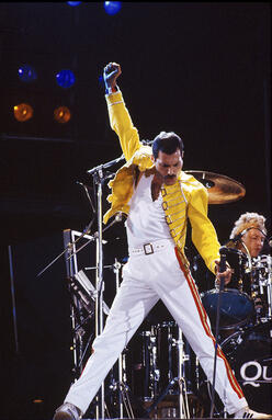 Singer Freddie Mercury wearing a yellow jacket performing on stage with his arm in the air 