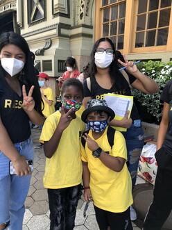 Two peer mentors and two lower school students smiling and posing with a peace sign. All four are wearing masks. The peer mentors wear a black rescue t-shirt and the two lower school students wear a yellow shirt with the IRC logo. 