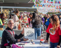 A World Bazaar volunteer poses with a man who holds up a piece of art. They are outside at the World Bazaar under flags from around the globe.
