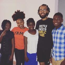 A man and a woman in their twenties, pose for a picture with three middle and high school age students in their home.