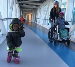 An intern pushing refugee child in a wheelchair in a hospital hallway, the other two siblings walking forward.