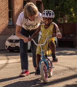 A man helping a refugee child ride a bike down the streets.