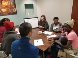 A group of refugees and tutors gather around a table with papers in front of them.