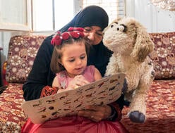 Ma'zooza, a baby goat Muppet on 'Ahlan Simsim', reads along with a young Syrian child and her caregiver in Azraq refugee camp in Jordan. 