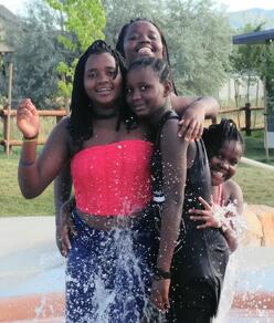 Four refugee girls stand in water park fountain smiling at the camera.
