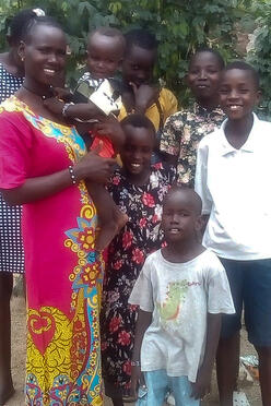 Florence Lokitoe poses with her children. They are standing outside and she is holding a baby, while five children stand next to her. 