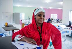 Dr Fatima poses with files at work wearing a red head scarf. There are hospital beds behind her. 