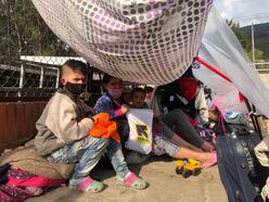 A mother with two kids sit under a makeshift tent surrounded by their luggage. They are all wearing masks and are looking directly at the camera. 
