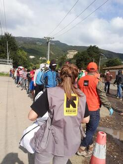 An IRC staff member stands with her back to the camera. In front of her is a line of people waiting to receive supplies. 