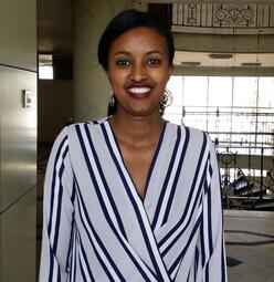 Betelhem Mengisu, wearing a black and white striped shirt and smiling at the camera, stands in a room near a spiral staircase. 