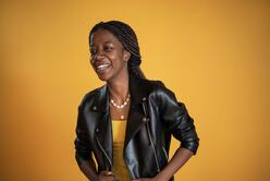Christelle, wearing a black leather jacket, smiles and poses in front of a yellow background. 